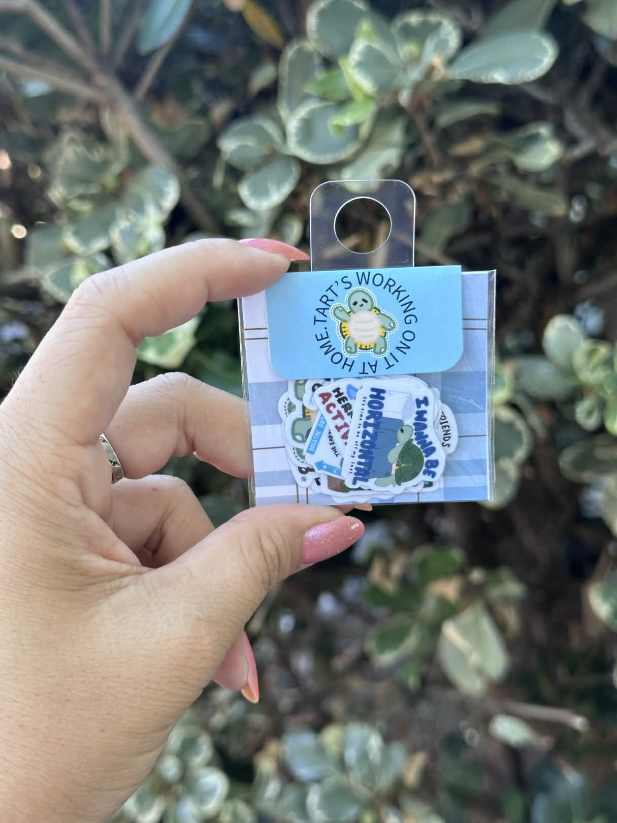 A hand holding a small packaged sticker set outdoors in front of green foliage. The package contains multiple colorful stickers, including a blue circular sticker that reads 'TART'S WORKING ON IT ALL THE TIME' and a white striped backing card.